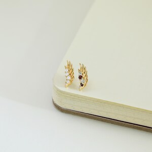 May include: A pair of gold-colored feather-shaped earrings. Each earring features a series of small, sparkling gemstones in white, red, and blue hues. The earrings are displayed on a cream-colored surface.