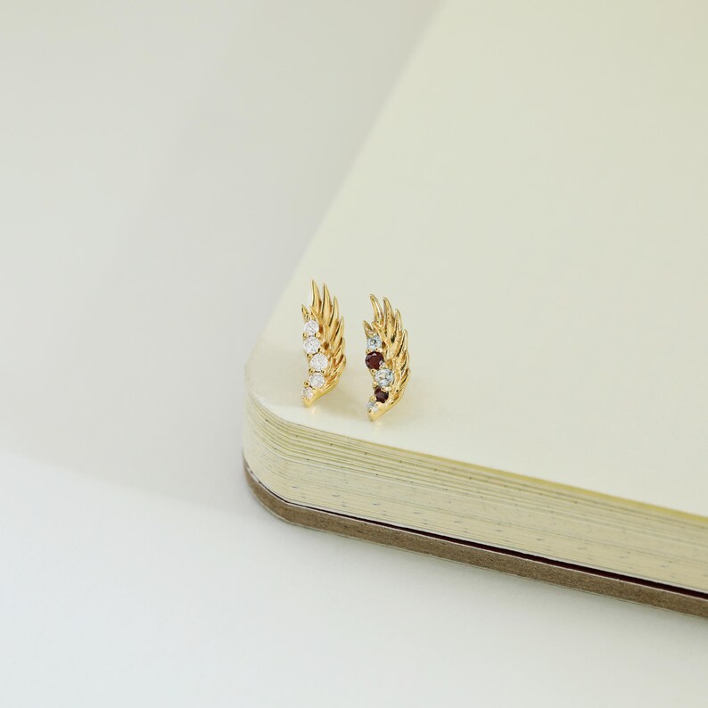 May include: A pair of gold-colored feather-shaped earrings. Each earring features a series of small, sparkling gemstones in white, red, and blue hues. The earrings are displayed on a cream-colored surface.