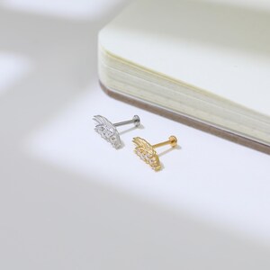 May include: Two small, decorative ear studs. One is silver-colored, the other is gold-colored. Each stud features a wing-like design with small, clear gemstones. The studs are displayed on a white surface.