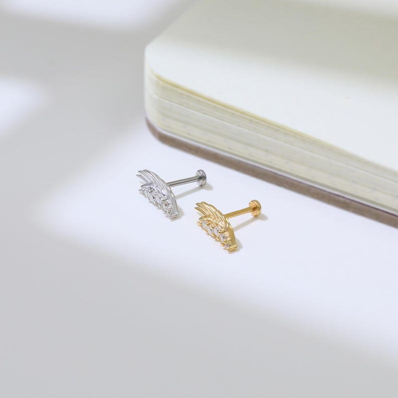 May include: Two small, decorative ear studs. One is silver-colored, the other is gold-colored. Each stud features a wing-like design with small, clear gemstones. The studs are displayed on a white surface.