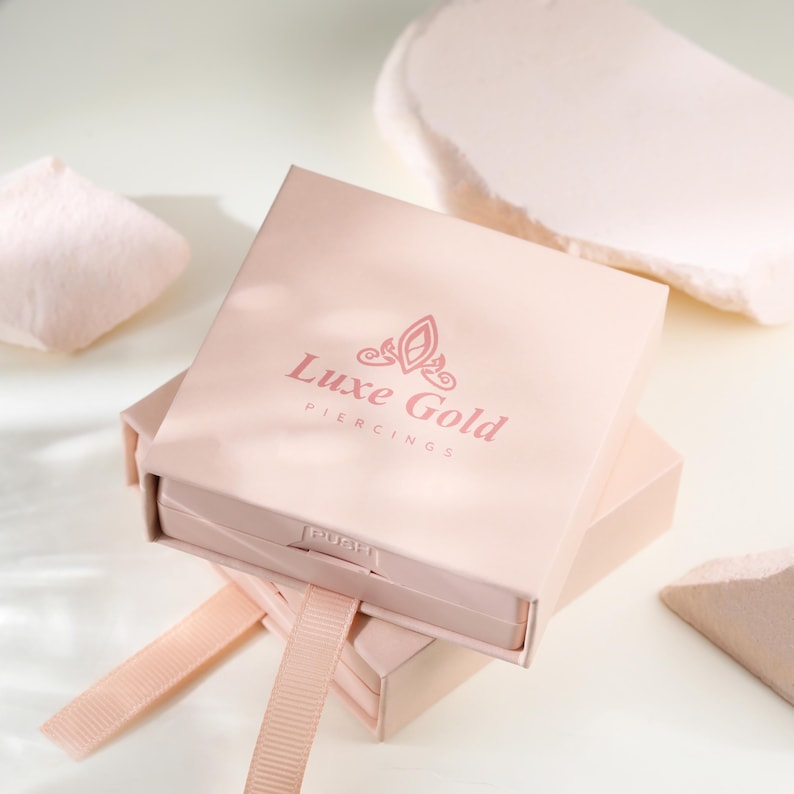 May include: A light pink box with the text "Luxe Gold Piercings" in a matching shade. The box is open, revealing a pull-out drawer with a ribbon. The background includes soft, abstract shapes in similar tones.