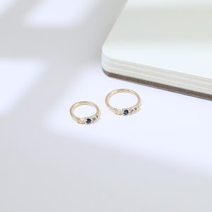 May include: Two gold rings, each featuring a small blue gemstone and two smaller clear stones. The rings are set against a white background with a notebook in the upper right corner. The rings have a delicate, elegant design.