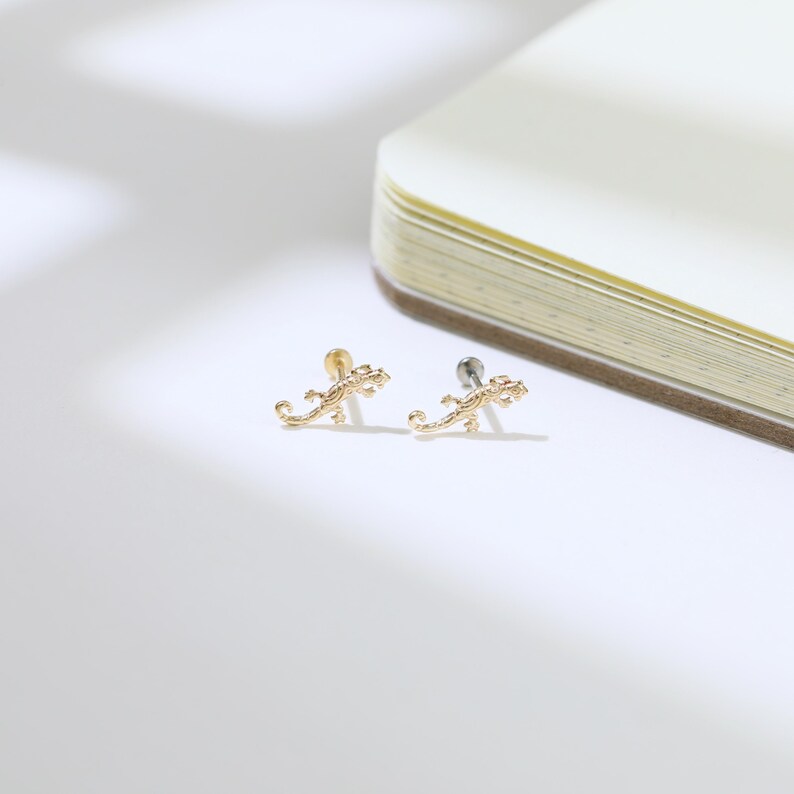 May include: Two gold-colored lizard-shaped stud earrings. The earrings are on a white surface next to a partially visible notebook. The earrings have a detailed design, with visible legs, tails, and heads. One earring has a gold post, and the other has a silver post.