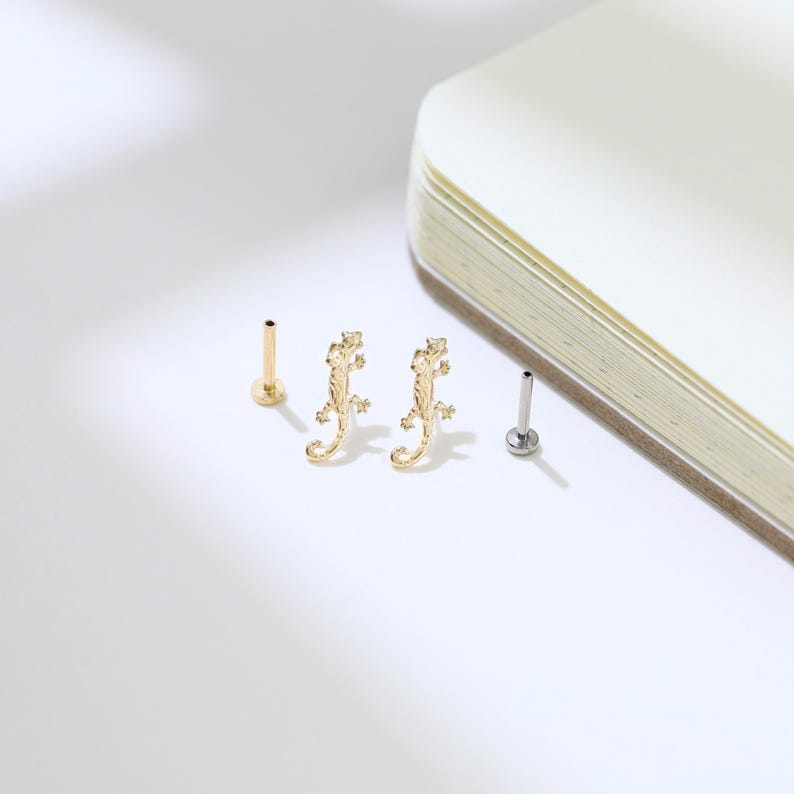 May include: Gold-colored lizard-shaped earrings and two stud earrings. The earrings are on a white surface next to a notebook. The lizard earrings have a detailed design, and the studs have a simple, minimalist look. Jewelry.