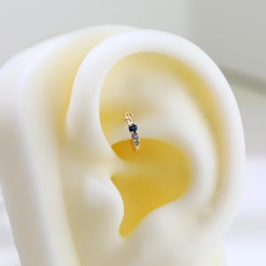 May include: A gold-coloured hoop earring featuring a line of small, dark blue and light-coloured gemstones. The earring is set against a light-coloured background, emphasising the jewellery's design. A delicate piece of jewellery.