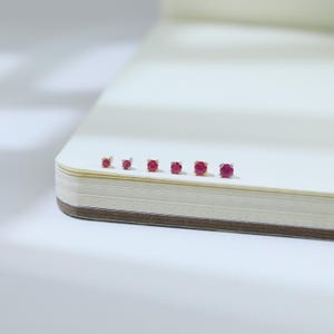 May include: A collection of six small stud earrings with round, vibrant red gemstones. The earrings are set in silver and gold-toned metal, displayed on a white surface. The gemstones are the focal point, showcasing their color and shape.