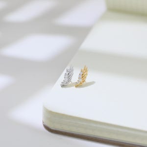 May include: Two stud earrings, one silver and one gold, each featuring a feather design. The silver earring has three clear stones, and the gold earring has three clear stones. The earrings are displayed on a white surface.