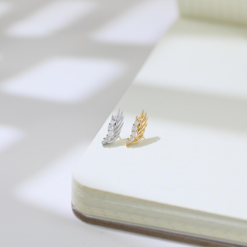 May include: Two stud earrings, one silver and one gold, each featuring a feather design. The silver earring has three clear stones, and the gold earring has three clear stones. The earrings are displayed on a white surface.