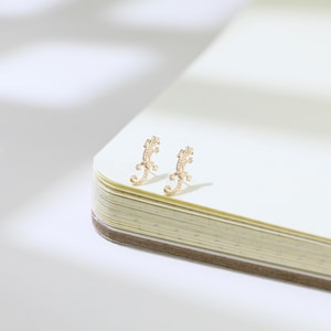 May include: A pair of gold-colored lizard-shaped stud earrings. The earrings are detailed with textured patterns and are positioned on the corner of an open notebook with cream-colored pages.