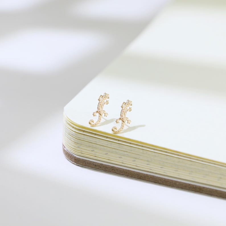 May include: A pair of gold-colored lizard-shaped stud earrings. The earrings are detailed with textured patterns and are positioned on the corner of an open notebook with cream-colored pages.