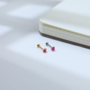 May include: Two small, stud earrings with a round, red gemstone. One earring has a gold post, and the other has a silver post. The earrings are displayed on a white surface next to a book.