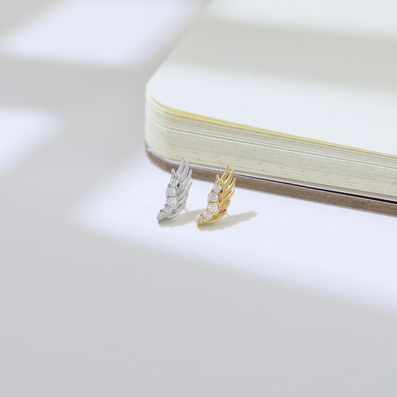 May include: Two wing-shaped earrings, one silver and one gold, each adorned with three clear, round gemstones. The earrings are set against a white background with a partially visible notebook.