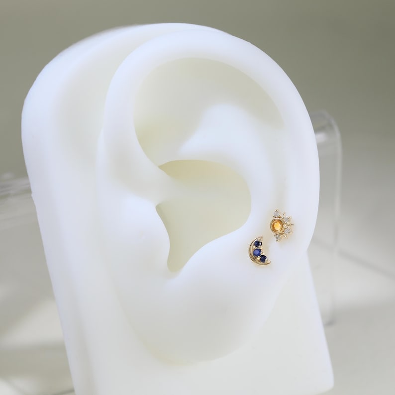 May include: A pair of gold earrings with small gemstones. The left earring is a crescent moon shape with three blue gemstones. The right earring is a starburst shape with a yellow gemstone in the center and smaller white gemstones around it.