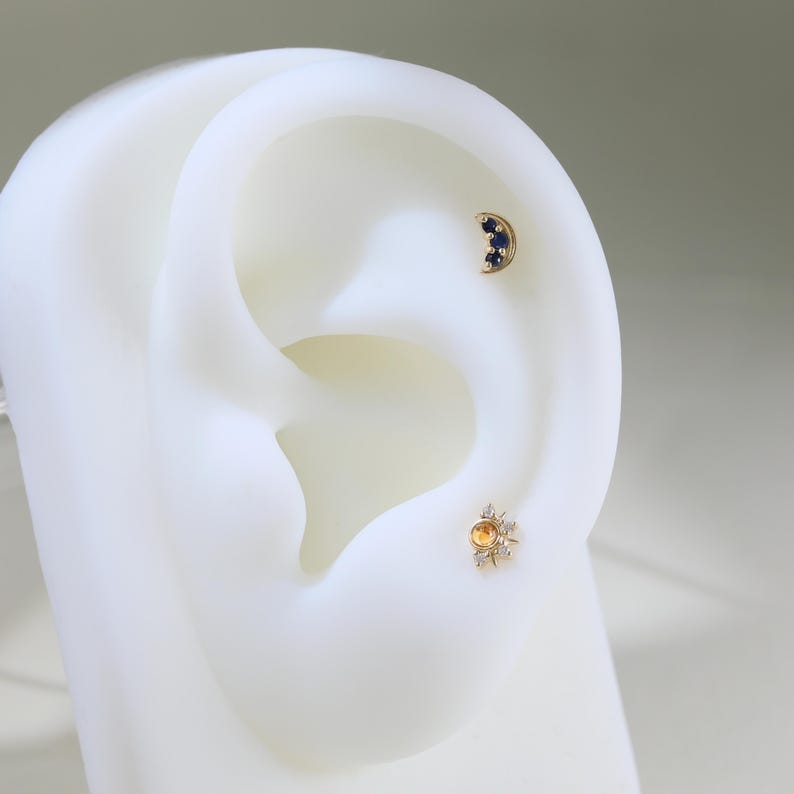 May include: A pair of gold earrings with gemstones. The left earring features a crescent moon shape with three blue sapphires. The right earring features a round yellow gemstone surrounded by smaller white diamonds.