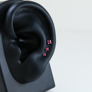 May include: Three small, square-cut ruby earrings displayed on a black ear model. The earrings are set in silver or white gold, with the vibrant red gemstones catching the light. The ear model is positioned against a white background.