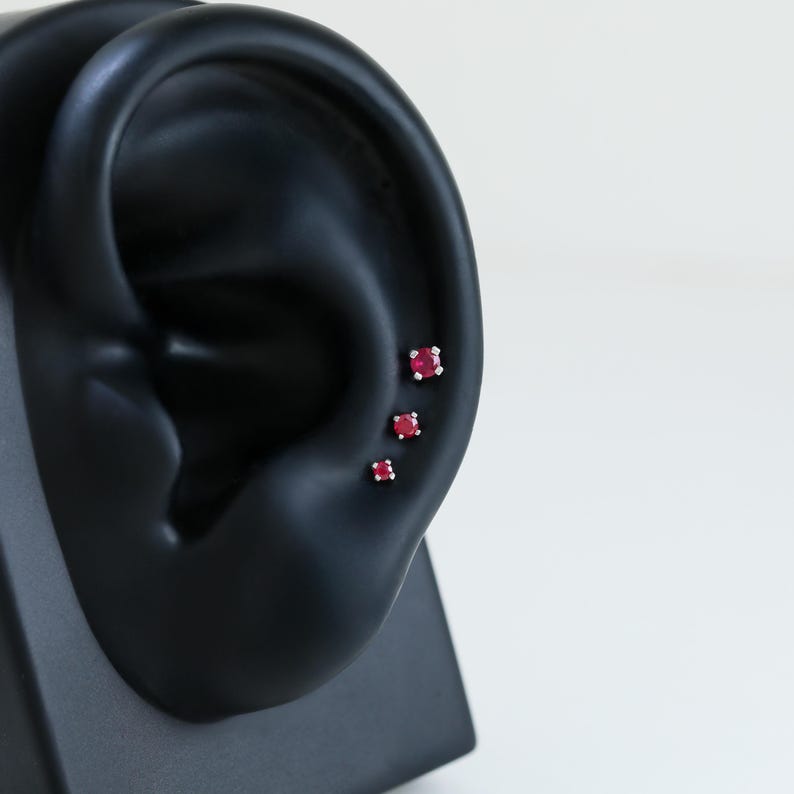 May include: Three small, square-cut ruby earrings displayed on a black ear model. The earrings are set in silver or white gold, with the vibrant red gemstones catching the light. The ear model is positioned against a white background.