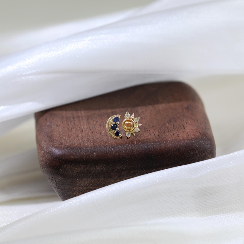 May include: A gold sun and moon earring with blue and white gemstones. The sun has a yellow center and the moon has a blue center.