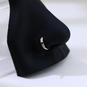 May include: A gold nose ring featuring a curved design. The ring is embellished with small, colourful gemstones, including blue, purple, and green. The jewellery is displayed on a black mannequin nose, against a white backdrop.