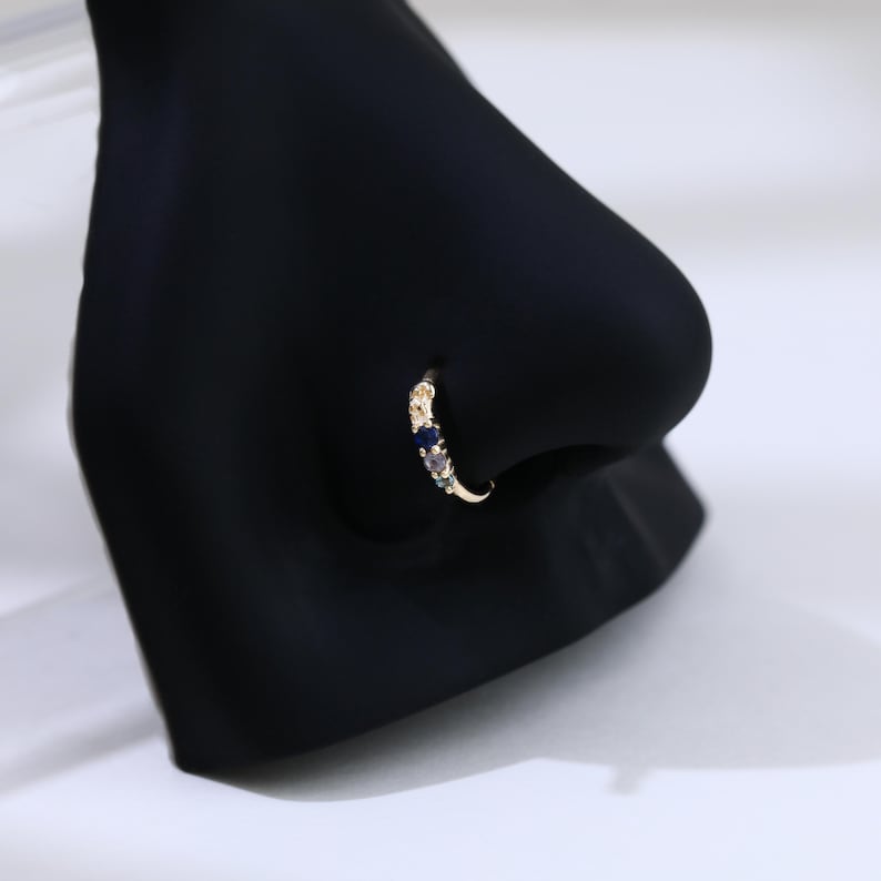 May include: A gold nose ring featuring a curved design. The ring is embellished with small, colourful gemstones, including blue, purple, and green. The jewellery is displayed on a black mannequin nose, against a white backdrop.