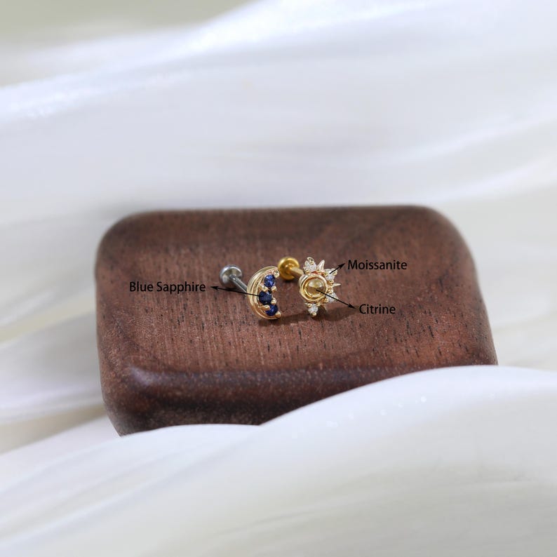May include: Gold moon and sun cartilage earring with blue sapphire and citrine gemstones. The sun features a center citrine stone surrounded by smaller clear stones, labeled "Moissanite".