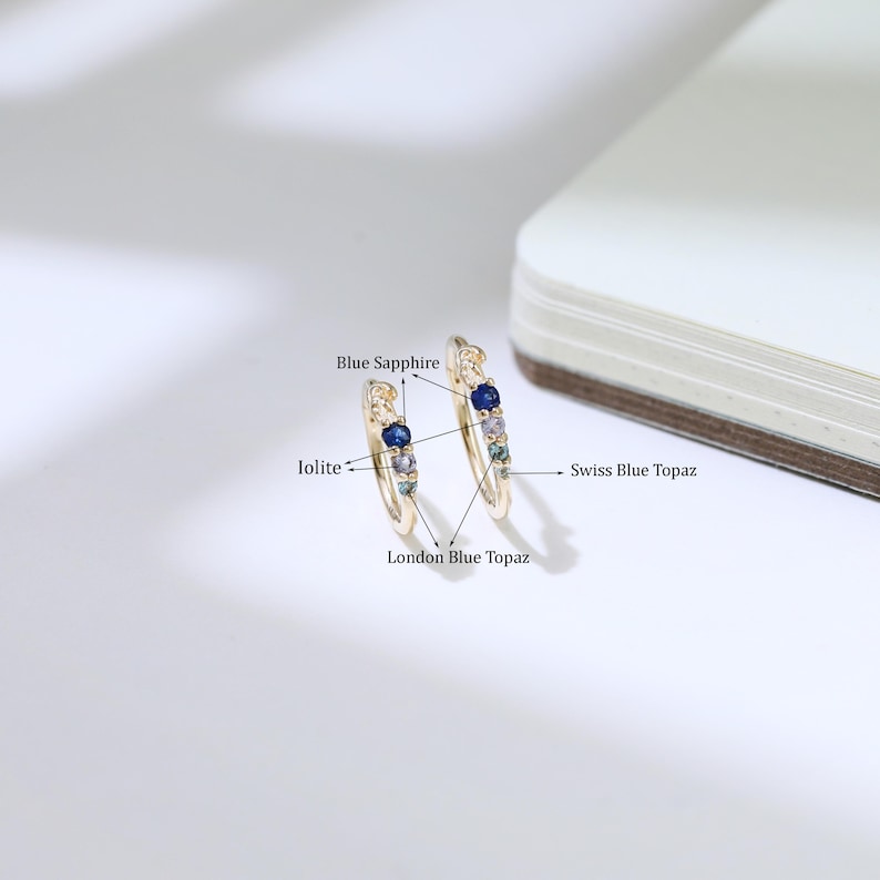 May include: A pair of gold hoop earrings, featuring blue sapphire, iolite, London blue topaz, and Swiss blue topaz gemstones. The earrings are presented on a white surface. The gemstones are in various shades of blue.