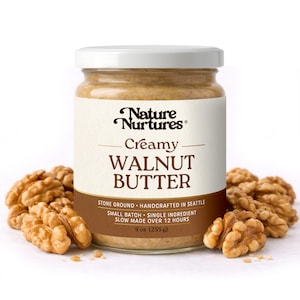 May include: A jar of creamy walnut butter with a white lid and a label that reads "Nature Nurtures Creamy Walnut Butter." The jar is surrounded by walnut halves. The label states the product is 9 oz (255 g).