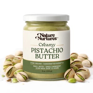 May include: A jar of creamy pistachio butter with a white lid and label. The label reads "Nature Nurtures Creamy Pistachio Butter." The jar is surrounded by pistachios in their shells. The jar contains 9 oz (255 g) of butter.