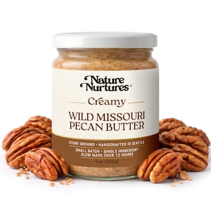 May include: A jar of creamy Wild Missouri Pecan Butter with a white lid and label. The label reads "Nature Nurtures" and "Creamy". The jar is surrounded by pecan halves. The jar contains 9 oz (255 g) of pecan butter.