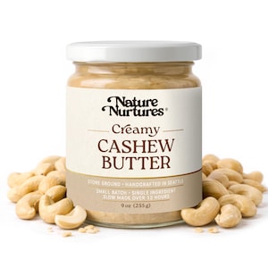 May include: A jar of creamy cashew butter with the text "Nature Nurtures Creamy Cashew Butter". The jar is surrounded by cashews. The label states the product is stone ground, handcrafted in Seattle, and contains 9 oz (255 g).
