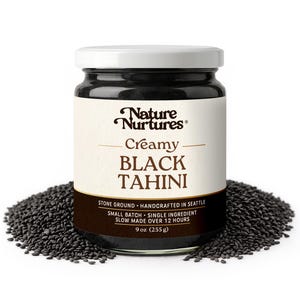 May include: A glass jar of creamy black tahini with a white lid, surrounded by black sesame seeds. The label reads "Nature Nurtures Creamy Black Tahini" and includes details about the product's origin and ingredients. The jar contains 9 oz (255 g).