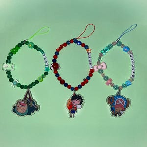 May include: Three beaded phone charms featuring anime character pendants. One green charm has a Zoro pendant, the second red and blue charm has a Luffy pendant, and the third charm has a Chopper pendant. Each charm has a unique colour scheme and bead style.