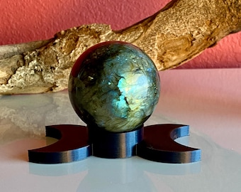 May include: A polished labradorite sphere with iridescent blue and green hues sits on a black crescent moon-shaped stand. The sphere reflects light, and a piece of driftwood rests in the background against a textured red wall.