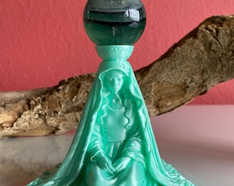 Może przedstawiać: Jasnozielona statua siedzącej kobiety w welonie, trzymającej ciemnozieloną kulę. Na podstawie statuy znajduje się napis "Lana Del Rey". Statua stoi na białej powierzchni z czerwonym tłem.