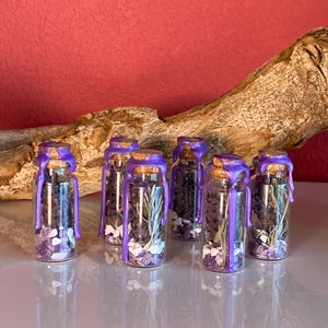 May include: Six small clear glass bottles with cork stoppers sealed with purple wax. Each bottle contains a mixture of dried lavender sprigs, small white stones, and purple amethyst crystals, creating a layered appearance inside the glass containers.