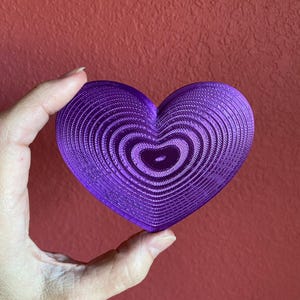 Może przedstawiać: Żywy fioletowy przedmiot w kształcie serca z koncentrycznym wzorem. Obiekt jest trzymany na czerwonym tle. Tekst "AMETHYST DRUZ" jest wyświetlany na dole obrazu.