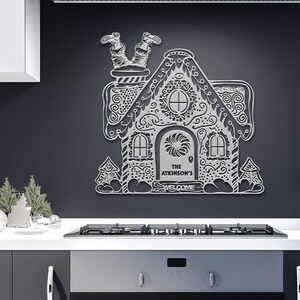 May include: A white gingerbread house wall decor with intricate details, including a chimney with a pair of legs sticking out, a wreath above the door, and the text "The Atkinson's Welcome".