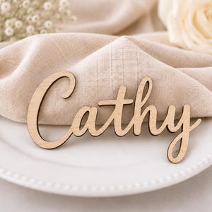 May include: A light-colored wooden cutout featuring the name Cathy in a cursive script font. The letters are connected in a single piece, showcasing the natural grain and texture of the wood material with a smooth, flat surface finish.