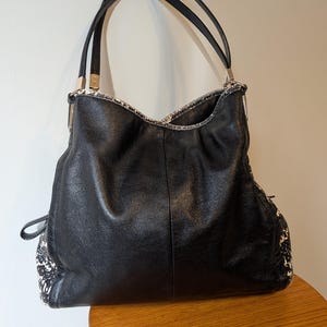 May include: A black leather hobo bag featuring a textured black and white snakeskin pattern on the side panels and trim. The handbag includes two thin black shoulder straps attached with silver-toned metal hardware and a central vertical seam.