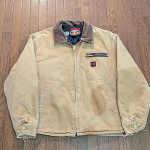 May include: A tan canvas work jacket with a brown corduroy collar and a front zip fastening. It features a black quilted lining, a zipped chest pocket, two lower hand pockets, and a black Tough Duck brand patch on the chest.