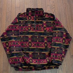 May include: A long-sleeved fleece pullover featuring a high collar with a partial front zip. The garment displays a repeating geometric pattern in shades of magenta, dark green, black, and yellow, with the word PENTAX embroidered in red.