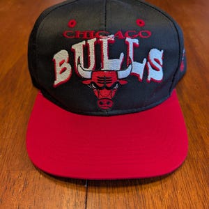 Puede incluir: Una gorra de béisbol negra con visera roja que presenta el texto bordado CHICAGO BULLS en letras blancas y rojas. El diseño incluye un logotipo central de un toro en rojo y blanco. La gorra tiene una corona estructurada y un botón rojo arriba.