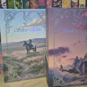 May include: Two hardback books with fantasy artwork. "Silver Elite" depicts a person on horseback in a landscape. "Broken Wings" shows a helicopter over mountains. Both have decorative borders and author names.
