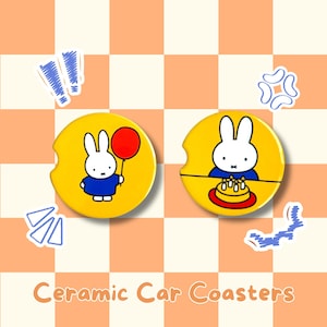 May include: Two yellow ceramic car coasters featuring a cartoon bunny character. One coaster shows the bunny holding a red balloon, the other shows the bunny with a cake. The text "Ceramic Car Coasters" is at the bottom.