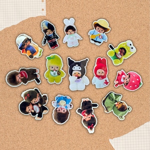 May include: A collection of colorful, cartoon-like stickers featuring various characters in different outfits and costumes. The stickers are arranged on a corkboard background, showcasing a variety of designs and themes, including animals and characters.