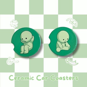 May include: Two green ceramic car coasters with a cartoon figure in different poses. The figure is light green with a simple design. The coasters have a cutout for easy removal and are set against a light green and white checkered background.