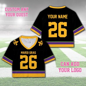 Pode incluir: Camisola preta com letras e acabamentos dourados. A camisola tem decote em V e apresenta o n&uacute;mero 26. O texto "MARDI GRAS" &eacute; exibido na frente. A camisola tamb&eacute;m tem o texto "YOUR NAME" nas costas. A camisola tem detalhes roxos e verdes.