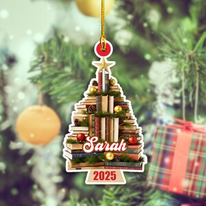 Può includere: Ornamento natalizio a forma di albero, composto da libri impilati, decorato con pigne, ornamenti rossi e dorati e una stella. Il nome "Sarah" è scritto in corsivo, con l'anno "2025" sotto.
