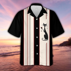 May include: A short-sleeve button-down shirt featuring a black collar and sleeves with a central cream-colored panel. The panel displays thin vertical red and black stripes and a black graphic of a cat holding a wine glass with star accents.