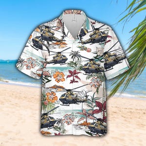 May include: White short-sleeved shirt with a tropical design featuring helicopters, palm trees, and flowers. The shirt has a classic collar and a relaxed fit, suitable for a casual look. The background shows a beach scene with blue water and sand.