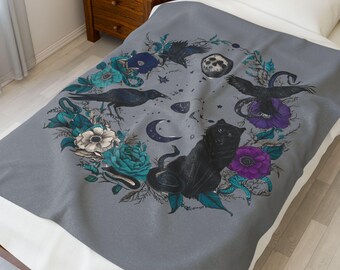 Puede incluir: Una manta gris con un diseño circular que presenta una pantera negra, cuervos, serpientes, flores y una luna creciente. El diseño incorpora elementos florales turquesa, morados y blancos. La manta está sobre una cama con una sábana blanca.